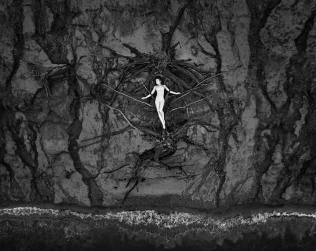 Entwined: The Figure as Landscape Black and white fine art photography made from drone by Tomas Rucker showing a central human figure framed by dark, skeletal tree roots against a rugged, erosion-patterned coast.